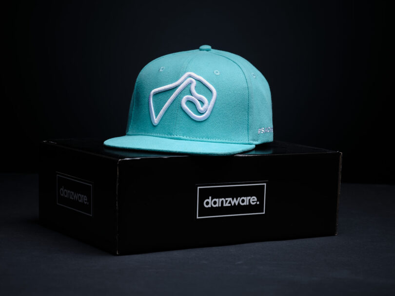 türkisfarbene Cap mit weißem sticke auf der Vorderseite. Der Stick ist in der Form der Sachsenringstrecke. Die Cap lieegt auf einem schwarzen Karton mit der Aufschrift "danzware". Der HIntergrund ist schwarz.