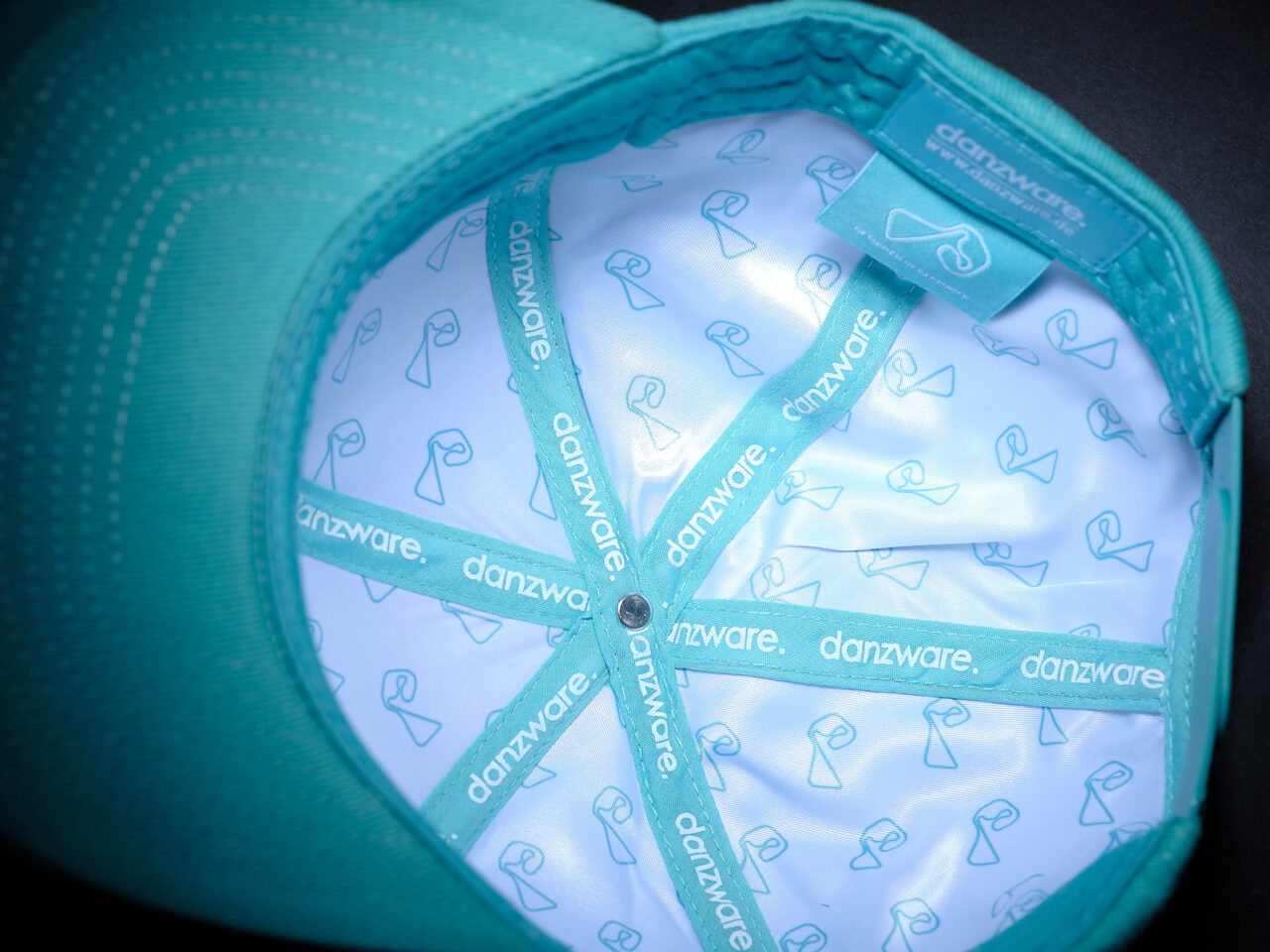 Innenansicht einer türkisfarbenen Kappe mit hellblauem Innenfutter, bedruckt mit dem Schriftzug 'danzware' und einem stilisierten Tanzsymbol.