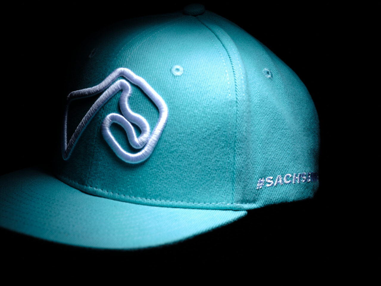 Türkisfarbenes Cap mit hochwertigem linienartigen Stick von der Sachsenring-Strecke und angeschnittener Aufschrift von 'SACHSENRING CIRCUIT' an der rechten Seite.
