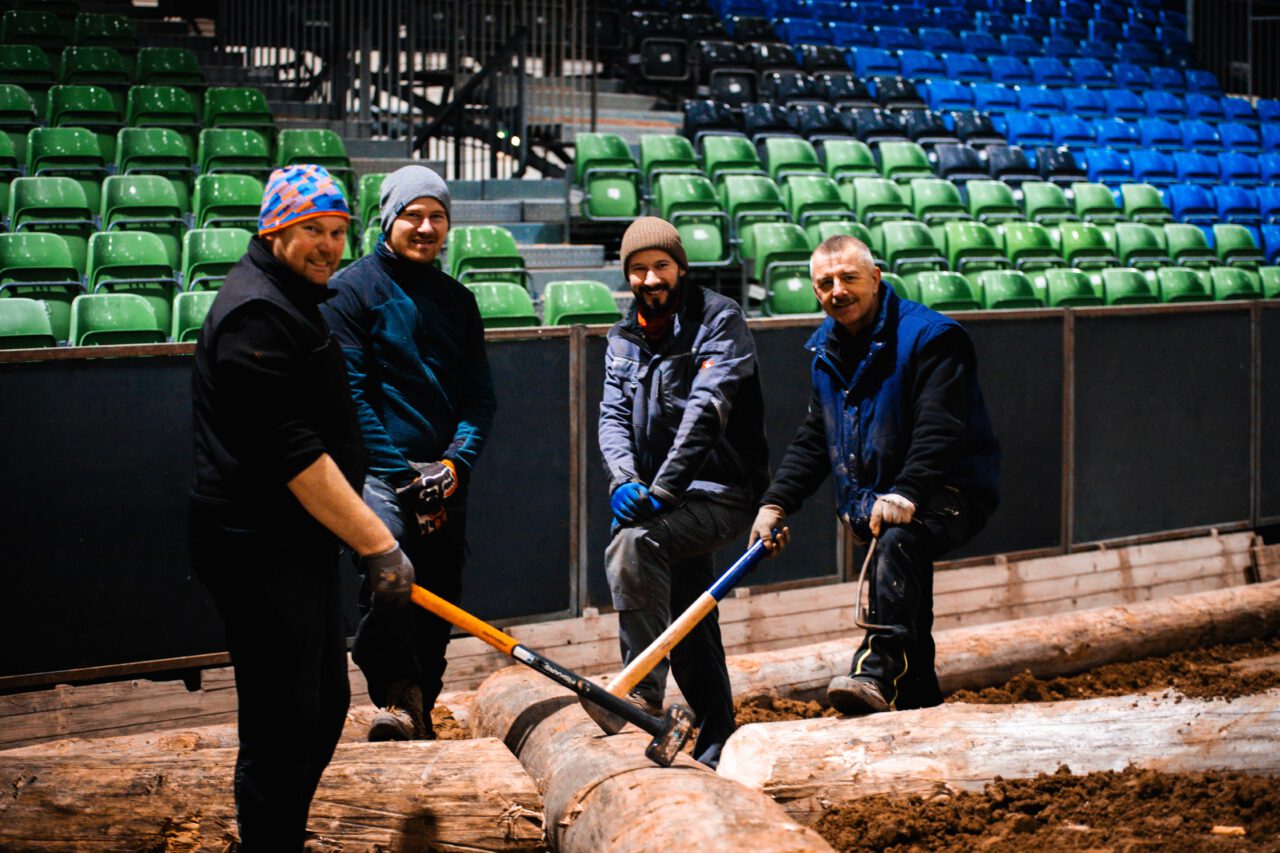 Vier Personen stehen auf einem Holzstamm in einer Arena mit grünen und blauen Sitzreihen, zwei halten Äxte zum Holzspalten.