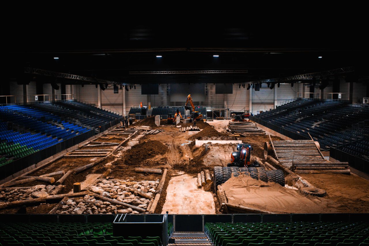 Große Halle mit Sitzreihen links und rechts, in der Mitte eine Baustelle mit Erde, Baumstämmen und Baumaschinen unter Deckenbeleuchtung.