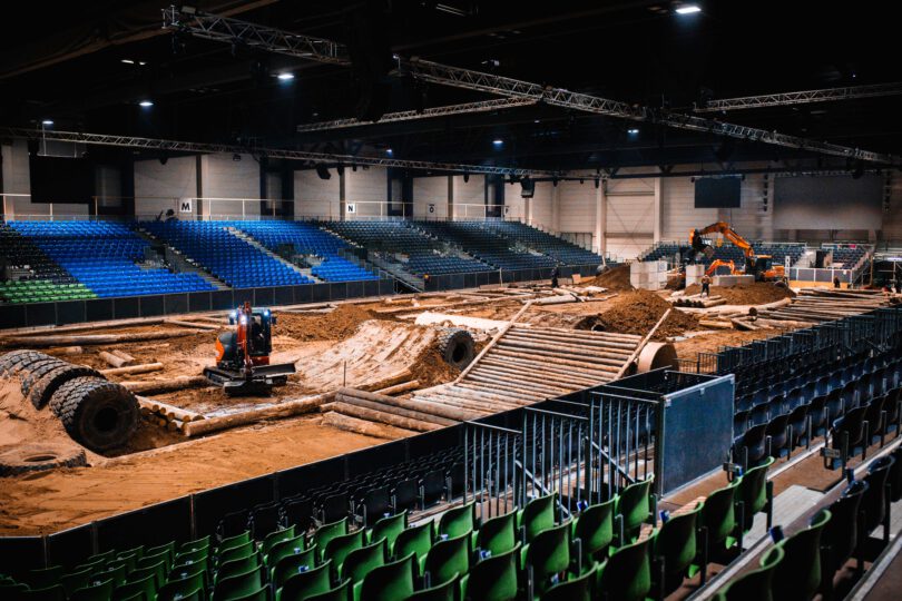 Leere Arena mit grünen und blauen Sitzreihen, im Innenraum Baustelle mit Erde, Rohren und zwei Baggern.
