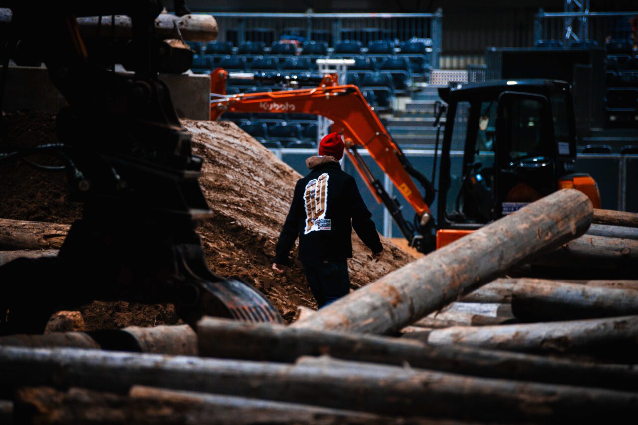 Person mit roter Mütze und schwarzem Pullover mit weißem Aufdruck geht zwischen Baumstämmen auf einer Baustelle mit Baggern und Erde.
