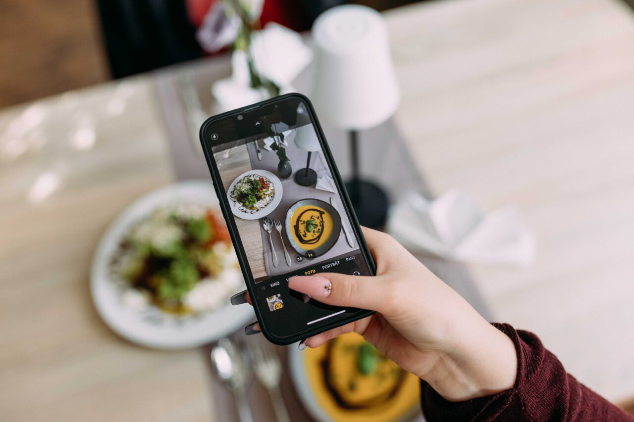 Bildschirm eines Handys wird gezeigt, der mit Kamerafunktion Essen fotografiert, was einer gelben Suppe und Salat entspricht, welches auf dem Tisch steht.