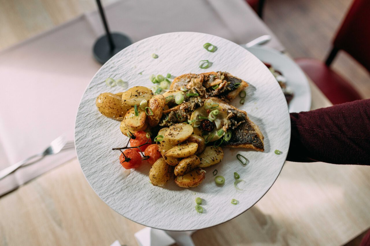 Gebratene Fischfilets mit gebackenen Kartoffeln, Kirschtomaten und Frühlingszwiebeln auf weißem Teller, gehalten von einer Hand.