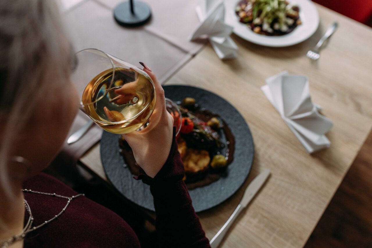 Frau mit blondem Haar hält ein Weinglas, vor ihr Teller mit Fleischgericht und Gemüse auf einem Holztisch mit gefalteten weißen Servietten.