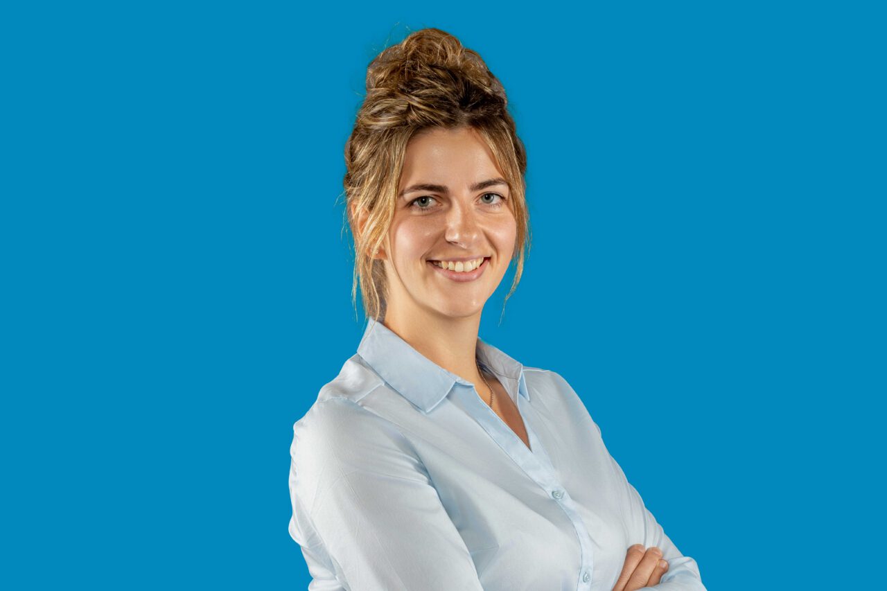 Junge hellhätuige Frau mit braunen Haaren zu einem dutt geschlossen, trägt helles Poloshirt und lächelt, sowei verschränkt die Arme