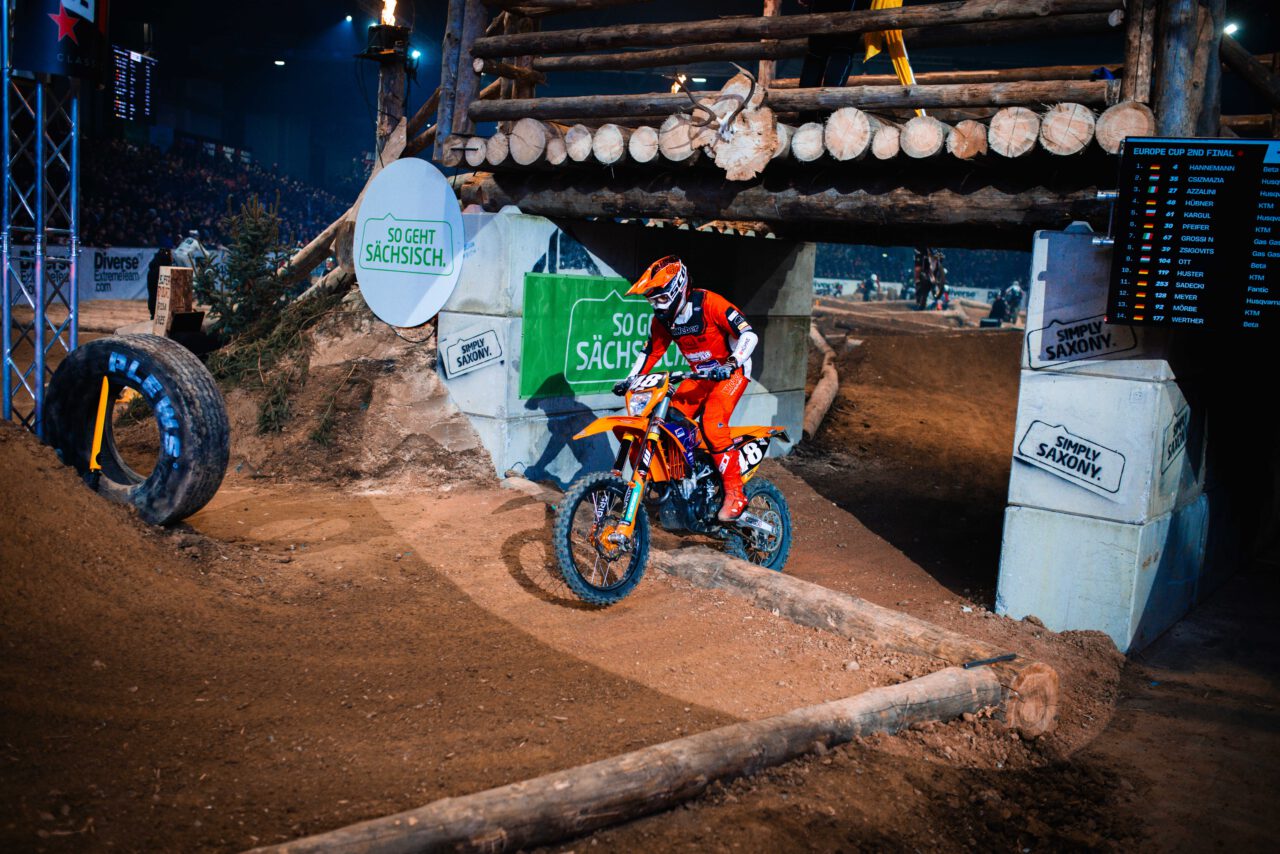 Motocross-Fahrer in orangefarbener Schutzkleidung und Helm fährt auf einem Dirtbike über eine Holzrampe in einer Indoor-Strecke mit Werbetafeln 'So geht Sächsisch'.