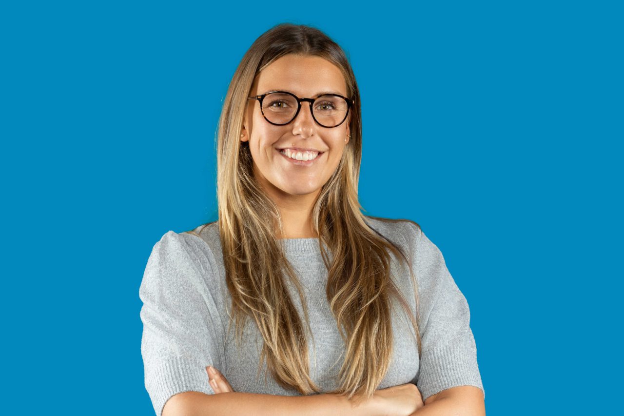 Junge hellhäutige Frau mit langen hellbrauenen Haaren verschränkt Arme, lächelt und trägt hellgraues Oberteil, sowie eine dunkelfarbene brille