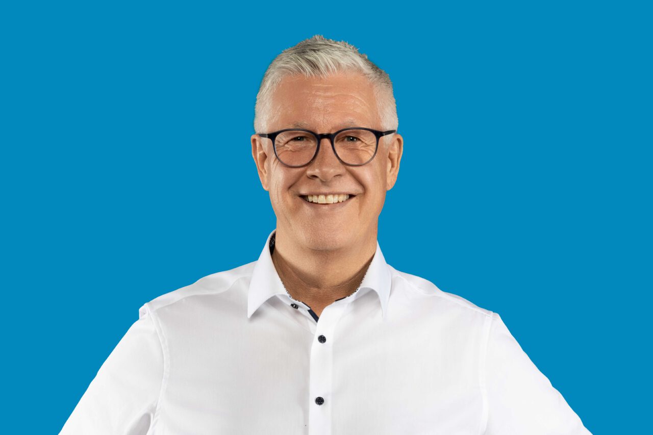 Mann mittleren Alters, hellhäutig, lächelt und trägt weißes Hemd, sowie eine schwarze Brille