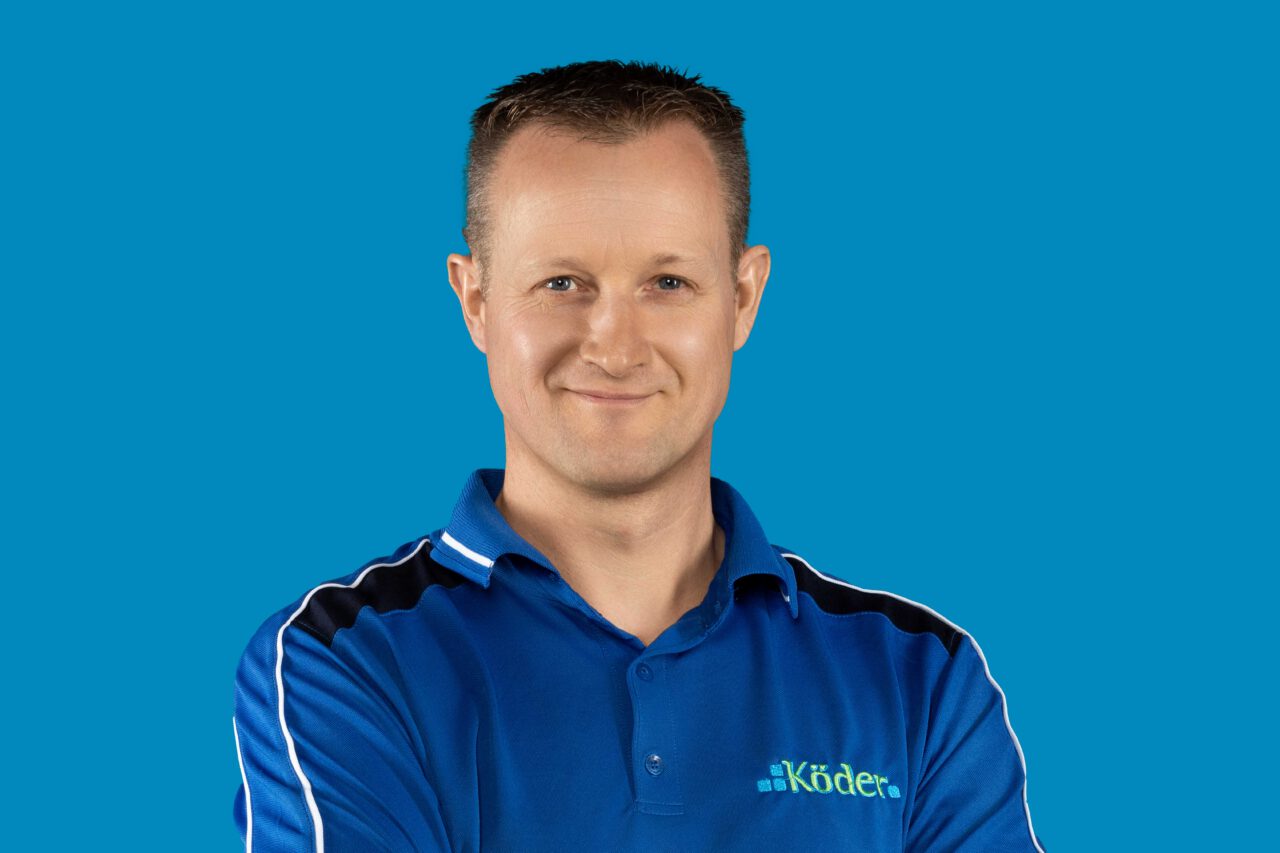 Mann mit schwarzen sehr kurzen Haaren, schmunzelndem Gesicht und dunkelblauer Jacke mit Aufschrfit "Köder" verschränkt Arme