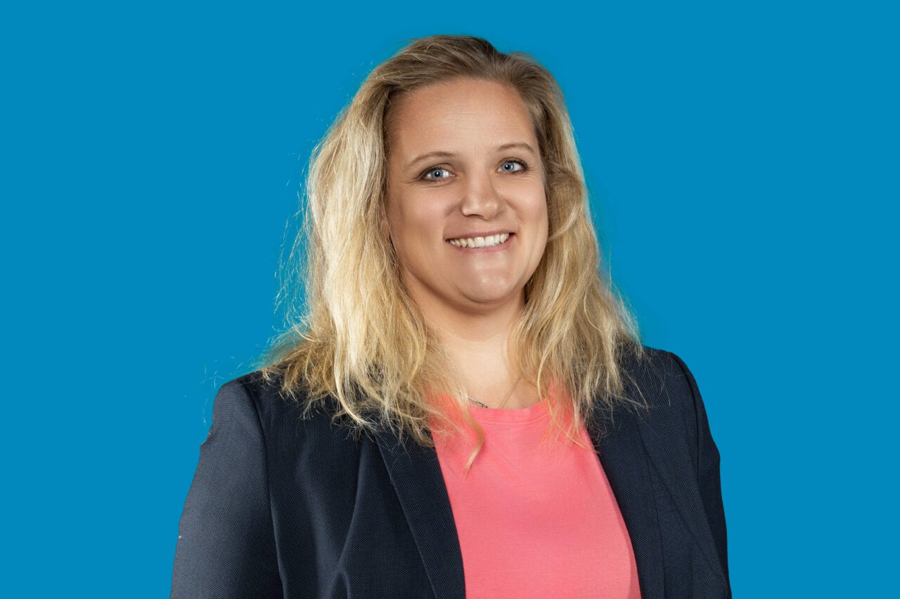 Hellhäutige Frau mit blonden Haaren trägt ein rosafarbenes Oberteil mit schwarzem Blaser und lächelt.