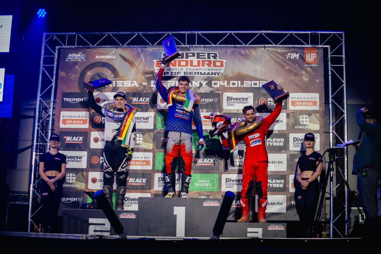 Drei Motorradrennfahrer in Rennanzügen stehen auf einem Podest mit den Plätzen 1, 2 und 3, halten Pokale hoch, umgeben von zwei Personen in schwarzer Kleidung und einem Fotografen.