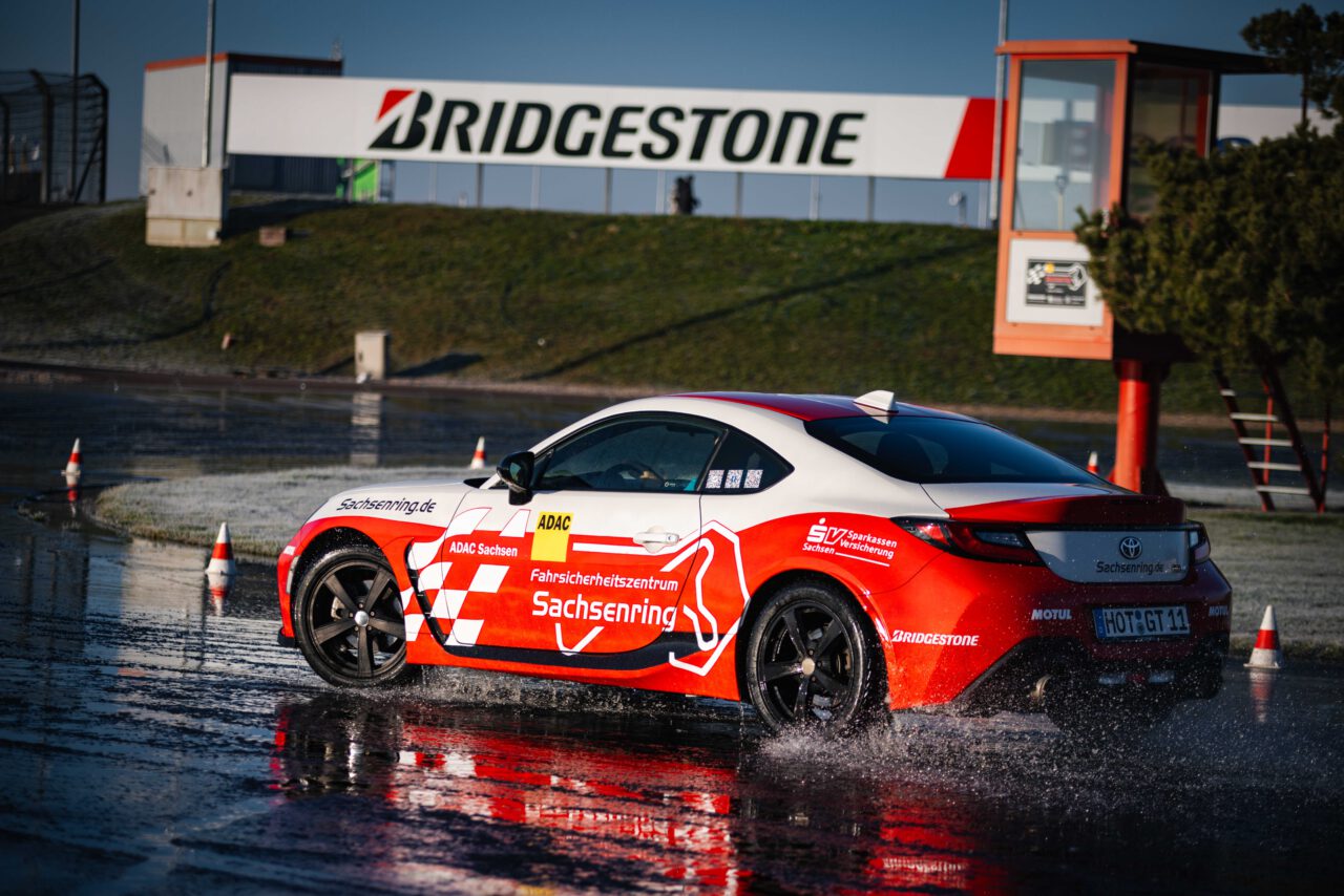 Roter und weißer Sportwagen mit Aufschrift 'Fahrsicherheitszentrum Sachsenring' fährt auf nasser Rennstrecke, Wasser spritzt seitlich hoch.