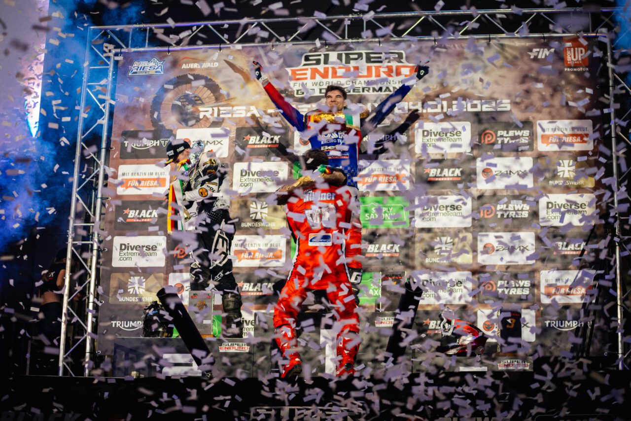 Drei Motorradrennfahrer in Rennanzügen stehen auf einem Podest mit den Plätzen 1, 2 und 3, halten Pokale hoch, umgeben von zwei Personen in schwarzer Kleidung, Konfetti und einem Fotografen.