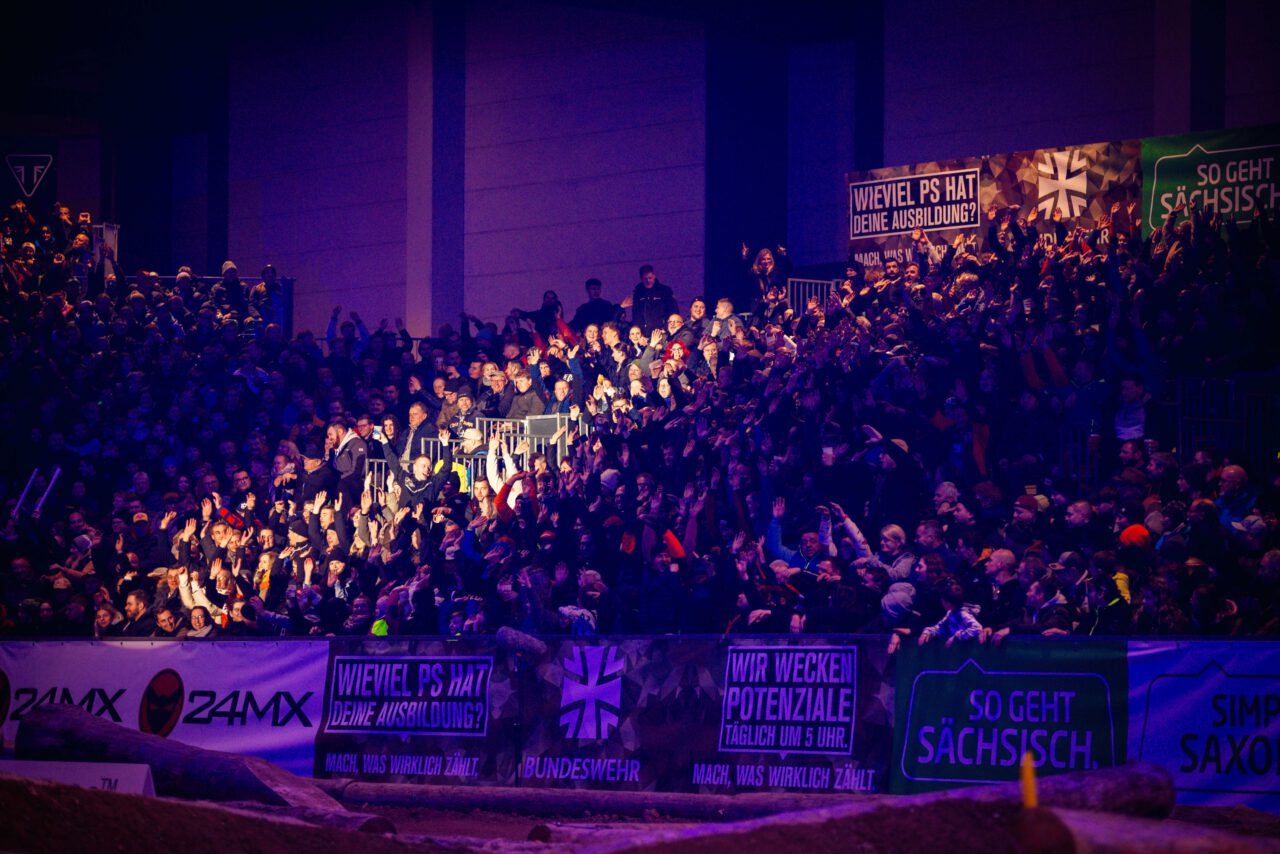Große Menschenmenge auf Tribüne bei Veranstaltung, viele halten Schilder mit Texten wie 'Wieviel PS hat deine Ausbildung?' und 'So geht sächsisch' hoch.