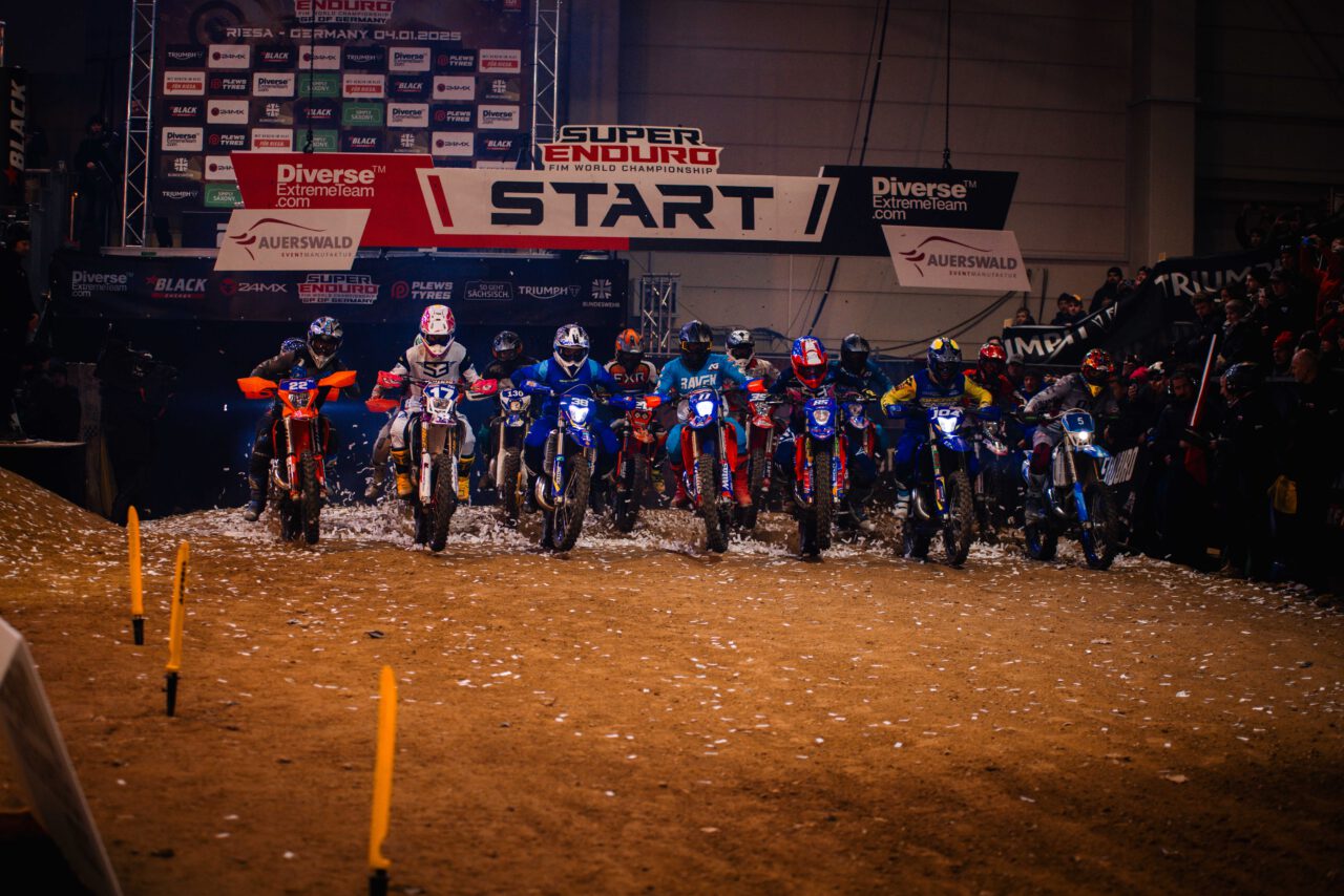 Sieben Motorradfahrer in voller Schutzkleidung starten ein Rennen auf einer Dirt-Strecke unter einem Banner mit der Aufschrift 'START'.