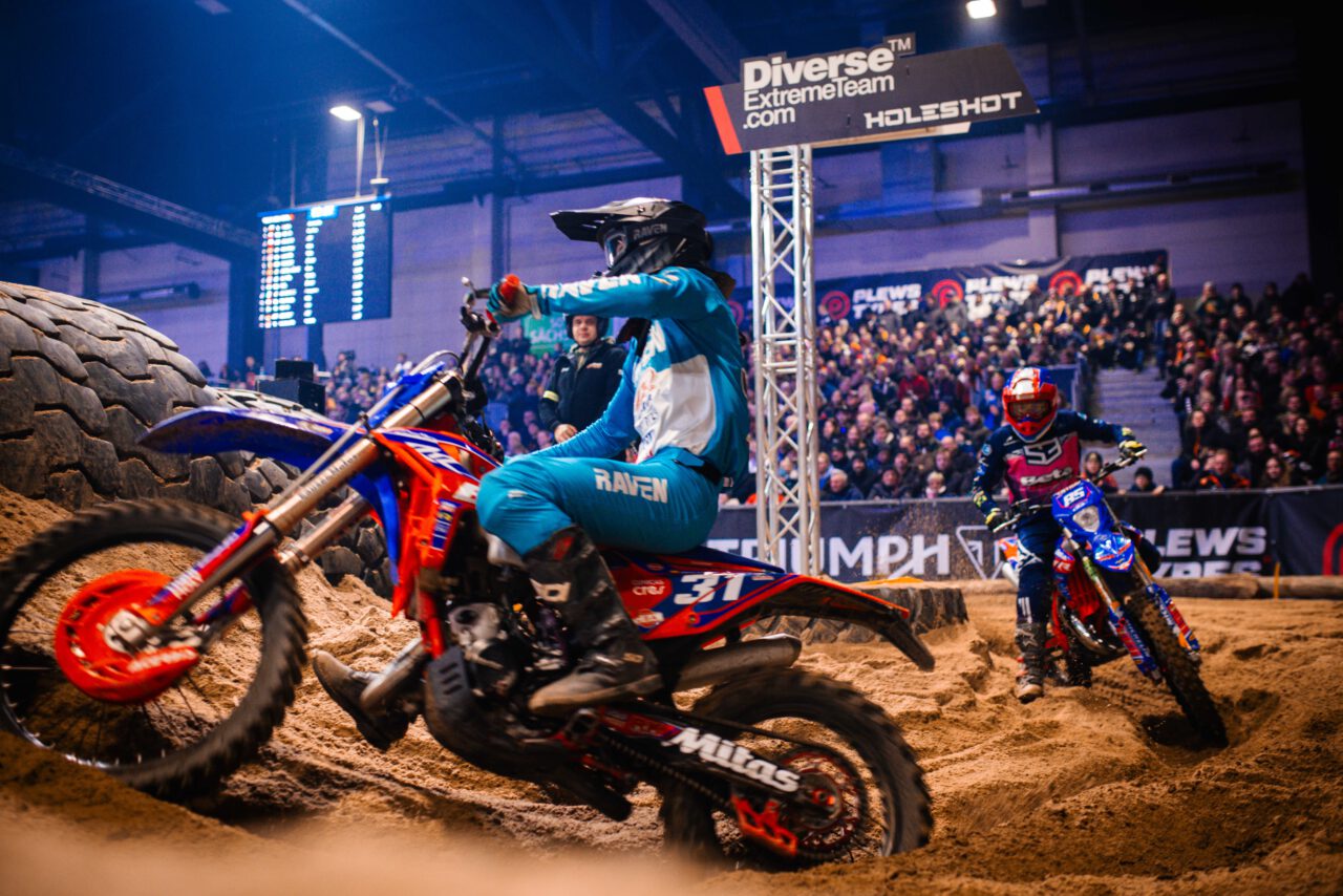 Motocrossfahrer in blauer Schutzkleidung auf orange-blauem Motorrad fährt auf sandigem Untergrund in einer Halle mit Publikum.