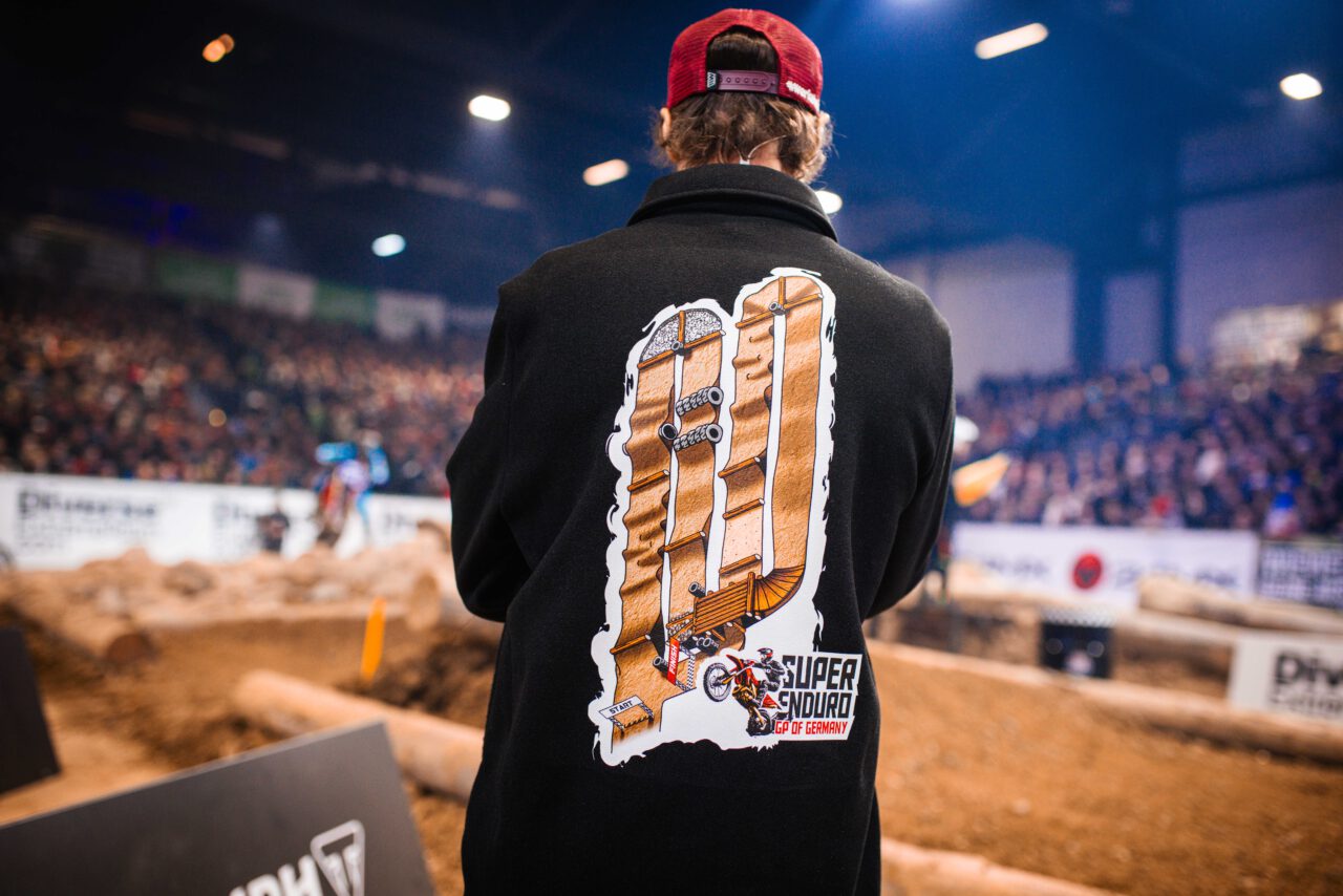 Rückansicht einer Person mit roter Kappe und schwarzem Mantel, auf dem ein großes, braunes Motorrad-Logo mit dem Schriftzug 'Super Enduro GP OF GERMANY' von ALROWA Designs zu sehen ist.