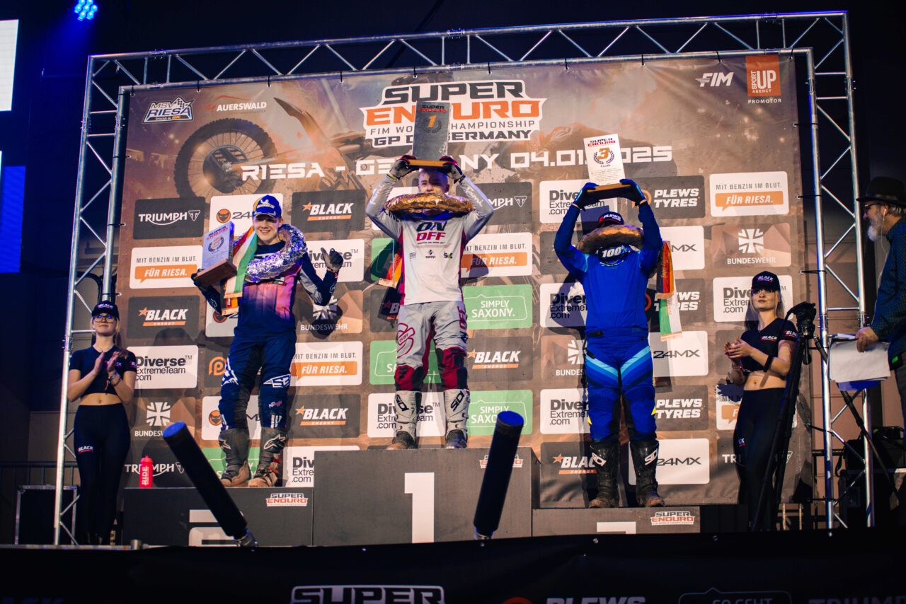 Drei Rennfahrer auf dem Siegerpodest, der mittlere trägt weiße Kleidung und hält eine Trophäe über dem Kopf, die anderen beiden stehen links und rechts in blauer Kleidung mit Trophäen in der Hand.