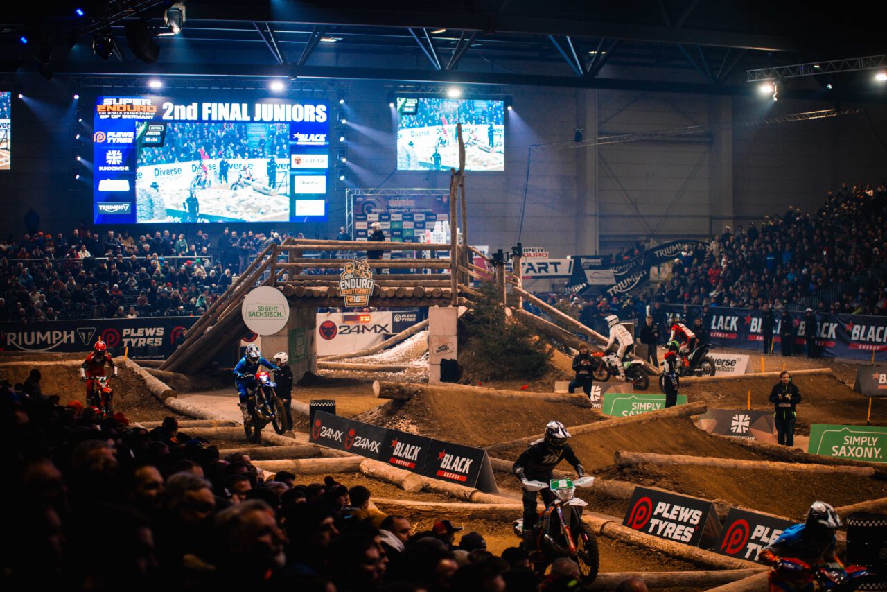 Motocross-Rennen in einer Halle mit mehreren Fahrern auf der Strecke und Zuschauern auf den Tribünen, große Anzeigetafel mit Text '2nd FINAL JUNIORS' im Hintergrund.