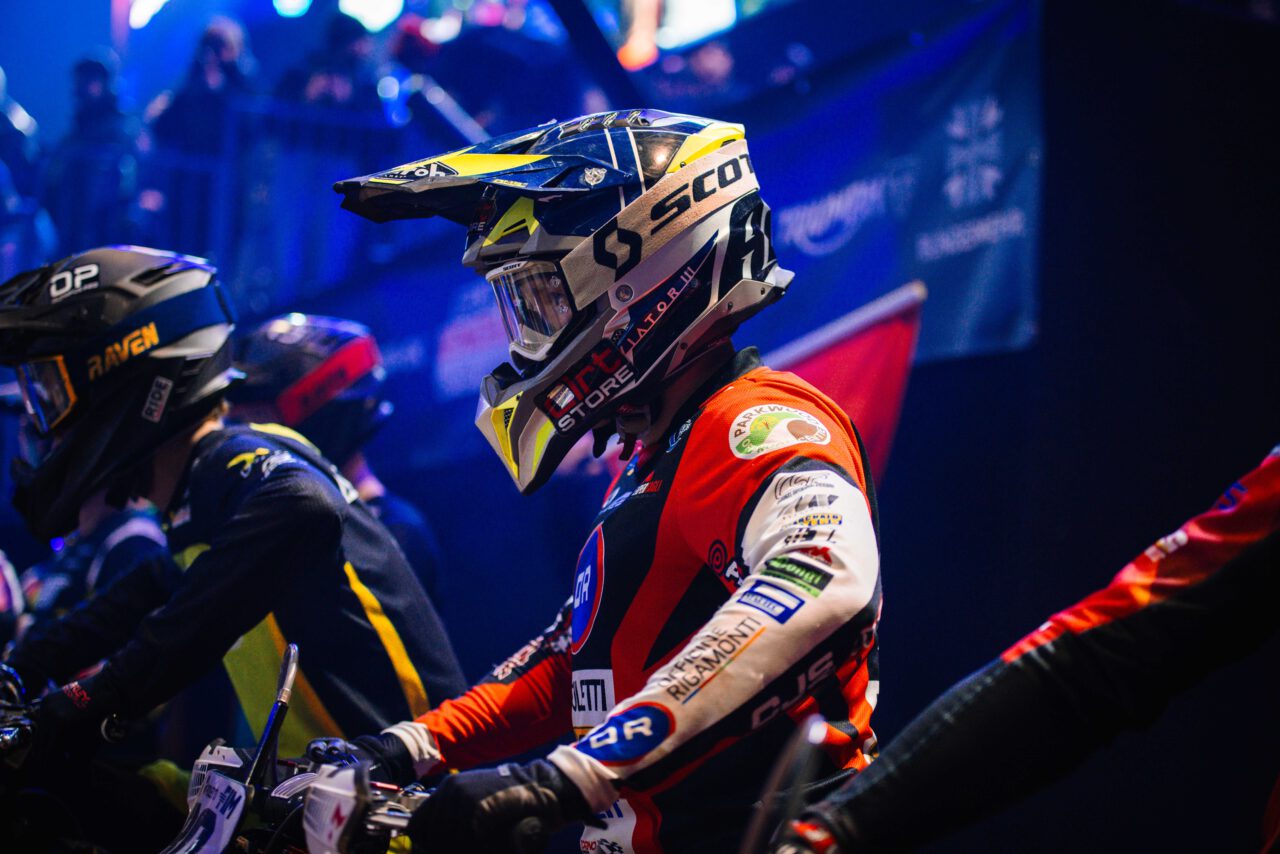 Motocross-Fahrer mit gelb-schwarzem Helm und rotem Trikot, bereit zum Start, umgeben von anderen Fahrern in dunkler Kleidung.