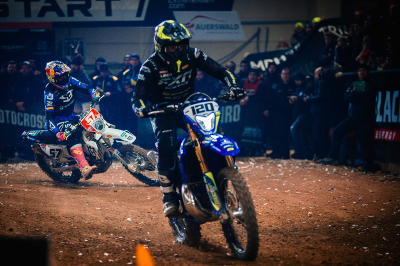 Zwei Motocross-Fahrer auf Rennstrecke, vorne Fahrer mit Nummer 120 auf blau-weißem Motorrad, dahinter Fahrer mit Nummer 57 auf weißem Motorrad.