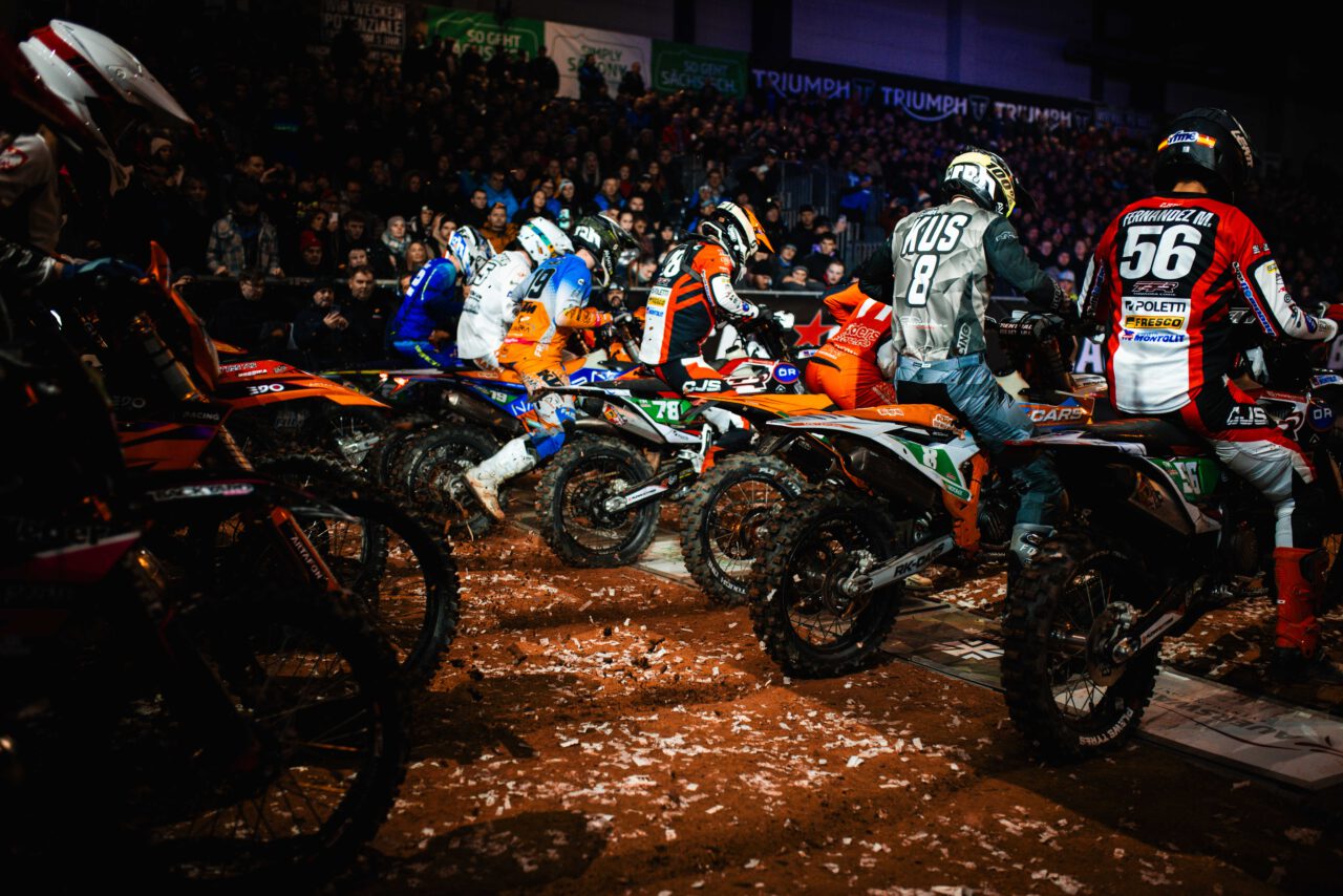 Mehrere Motocross-Fahrer in bunter Schutzkleidung und Helmen starten auf schlammigen Motorrädern bei einem Rennen in einer Halle.