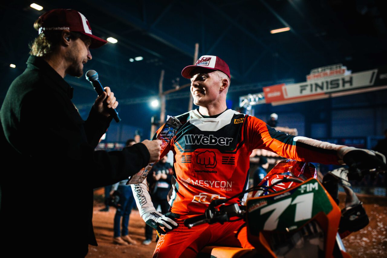 Motocross-Fahrer in rotem Anzug und weiß-roter Kappe mit Startnummer 71 auf dem Motorrad, spricht mit einem Mann mit Mikrofon in einer Halle mit Rennstrecke.