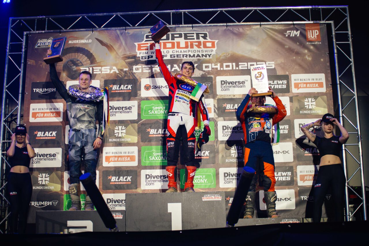 Drei Motorradfahrer in Rennanzügen stehen auf einem Podest mit den Plätzen 1 bis 3, halten Pokale hoch, im Hintergrund Banner mit Sponsoren und Veranstaltungstitel 'Super Enduro FIM World Championship'.