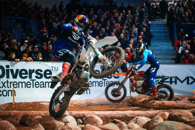 Motocrossfahrer in blauer Schutzkleidung springt mit weißem Motorrad über Felsen, im Hintergrund ein weiterer Fahrer auf rotem Motorrad und Zuschauertribüne