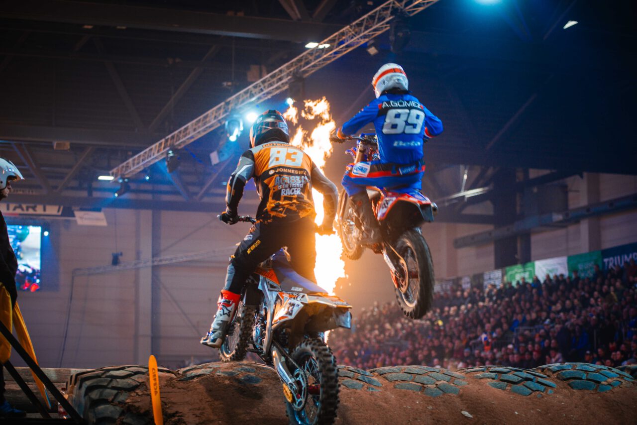 Zwei Motocross-Fahrer springen mit ihren Motorrädern über große Reifen in einer Halle vor Publikum.