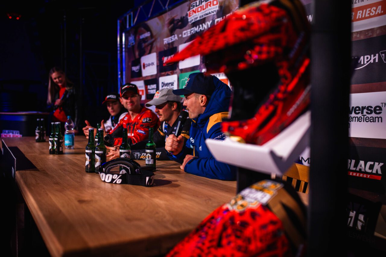 Motorradfahrer in Rennkleidung sitzen an einem langen Tisch mit Helmen und Getränkeflaschen, im Hintergrund Werbebanner.