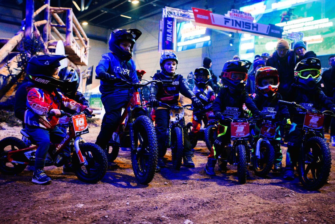 Mehrere Kinder mit Helmen auf kleinen Fahrrädern und Motorbikes stehen auf einer Dirt-Strecke vor einem Zielbogen mit der Aufschrift 'FINISH'.