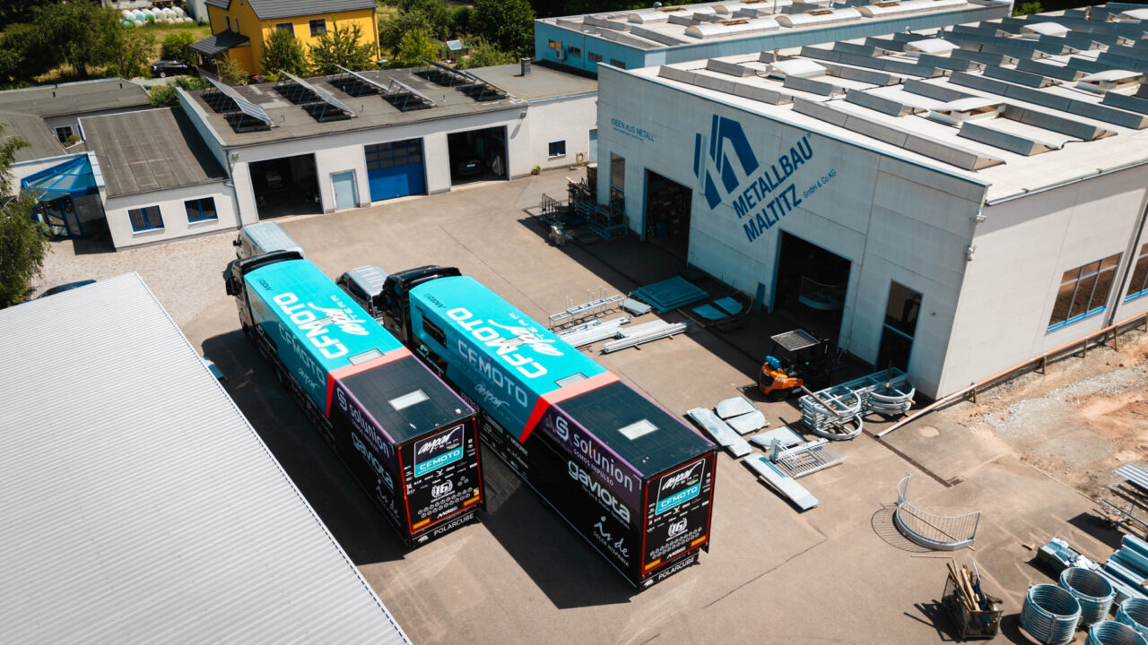 Luftaufnahme von zwei schwarzen Lkw-Anhängern mit türkisfarbenen Planen vor einem Gebäude mit dem Schriftzug 'Metallbau Maltitz'.