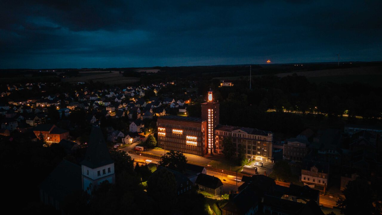 Nachtaufnahme einer beleuchteten Stadt mit einem hohen, hell erleuchteten Turm und umliegenden Häusern unter bewölktem Himmel.