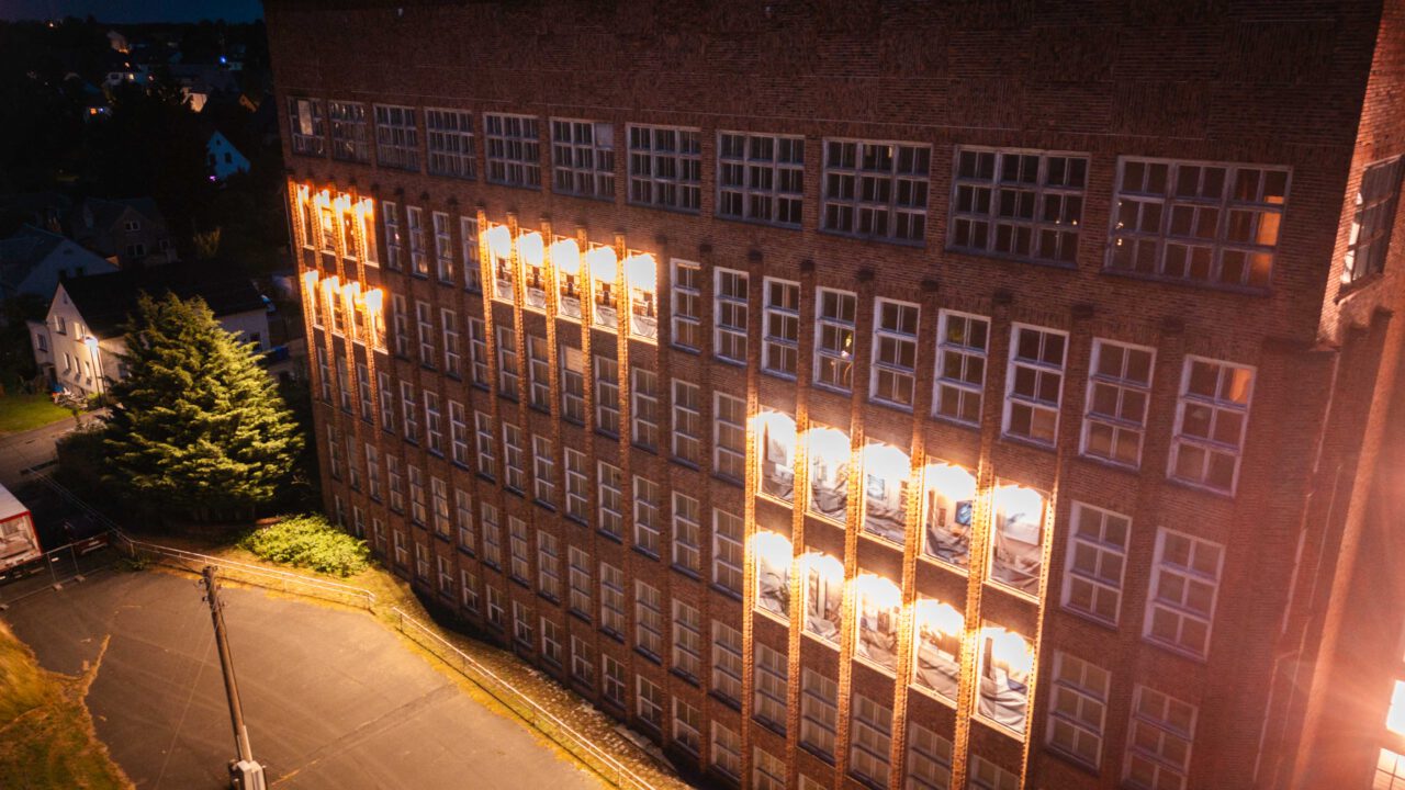 Beleuchtete Backsteinfassade eines mehrstöckigen Gebäudes mit vielen Fenstern bei Nacht.