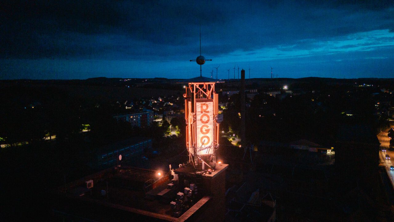 Beleuchteter Turm mit dem Schriftzug 'ROGO' in einer nächtlichen Stadtlandschaft mit Windrädern am Horizont.