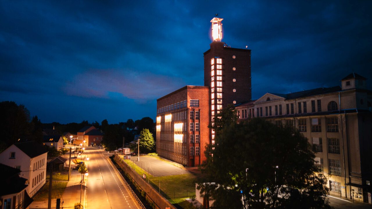 Beleuchtetes Industriegebäude mit Turm und rotem Leuchtsymbol bei Nacht an einer leeren Straße.