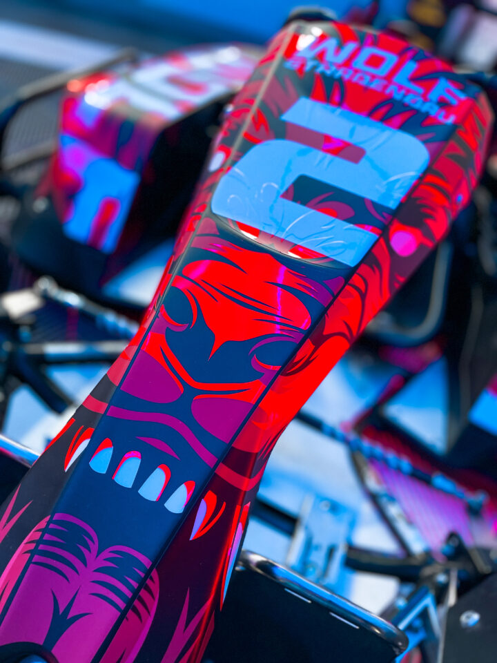 Nahaufnahme des Cockpitbereichs eines Rennwagens mit auffälligem rot-blauem Wolfkopf-Design und der Zahl '2' in weiß im blauen Neonlicht.