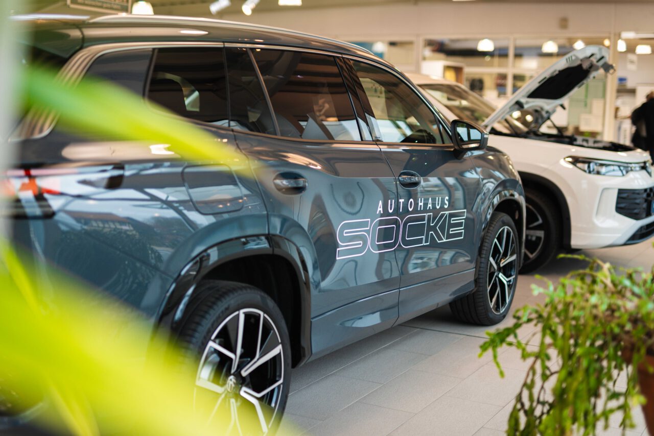 Seitenansicht eines grauen Autos mit der Aufschrift 'Autohaus Socke' auf der Tür, im Hintergrund ein weiteres Auto mit geöffneter Motorhaube in einem Ausstellungsraum.