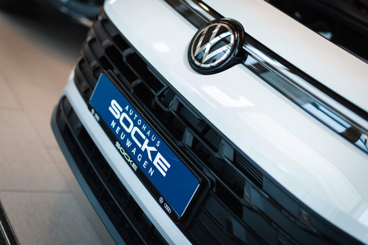 Nahaufnahme des Kühlergrills eines weißen Volkswagen mit blauem Nummernschild 'Autohaus Socke Neuwagen'