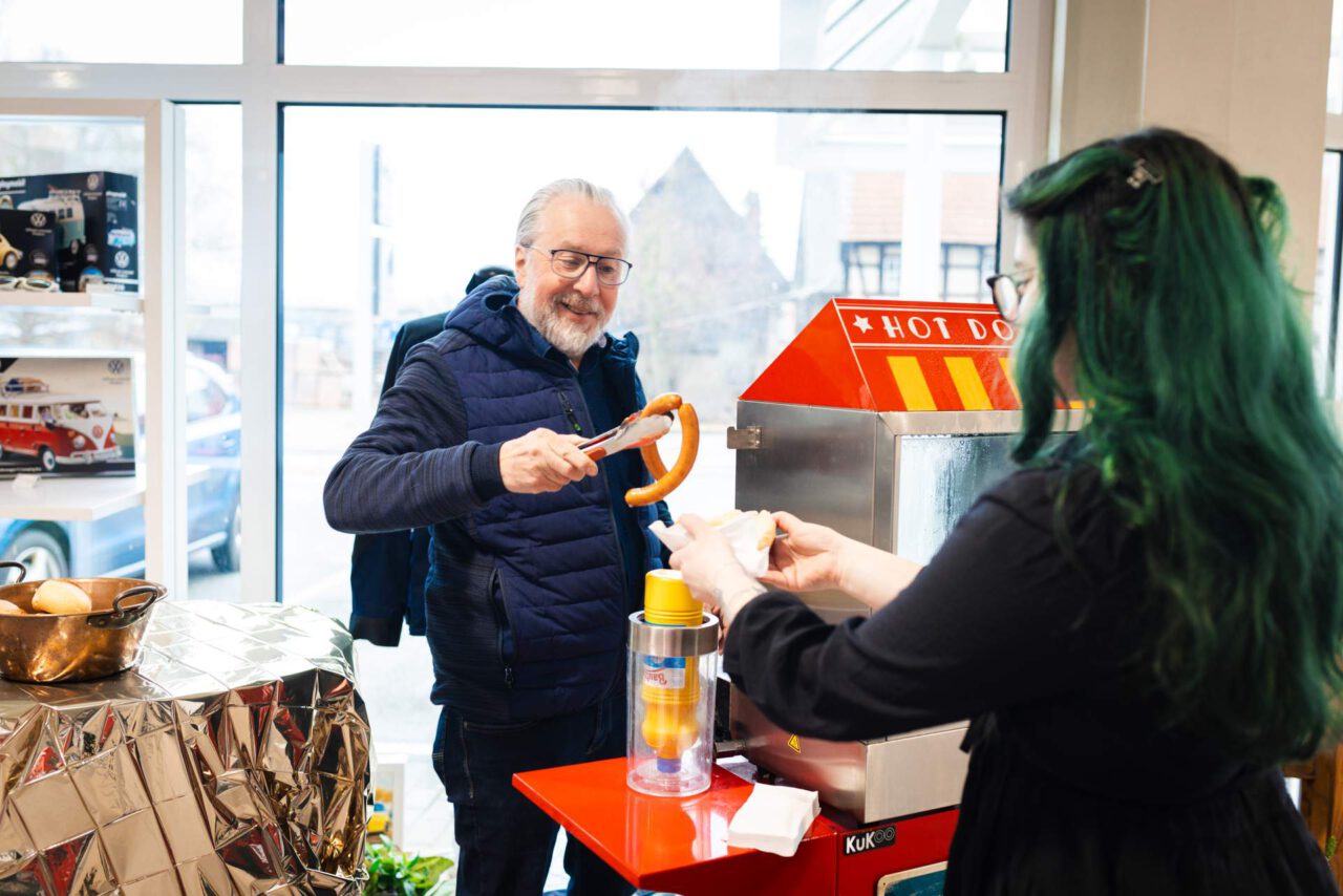 Mann in blauer Jacke nimmt mit Zange eine Bratwurst vom Hot-Dog-Stand, Frau mit grünen Haaren reicht Brötchen.