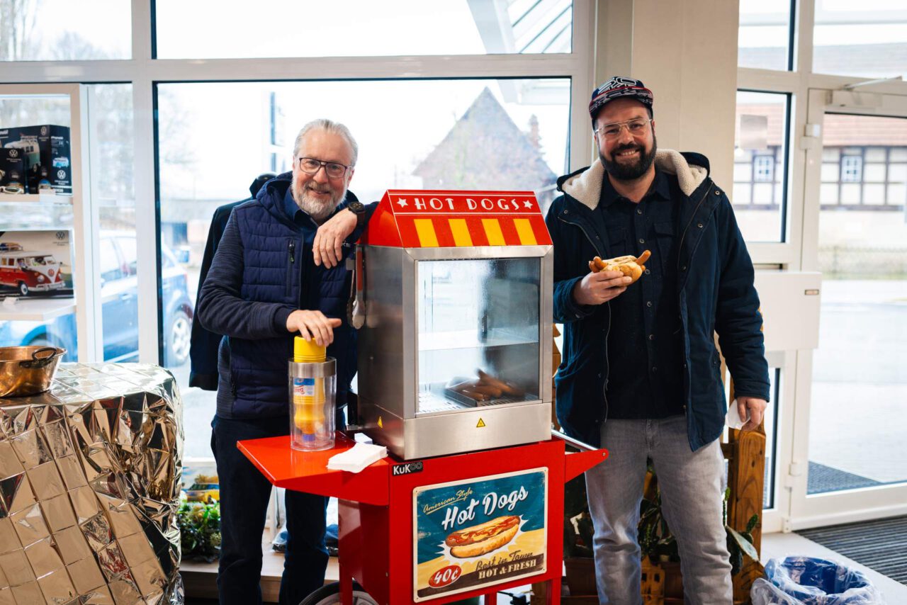Zwei Personen stehen neben einem roten Hot-Dog-Stand mit Glasvitrine und Schriftzug 'Hot Dogs'. Eine Person hält einen Hot Dog, die andere eine Getränkedose.