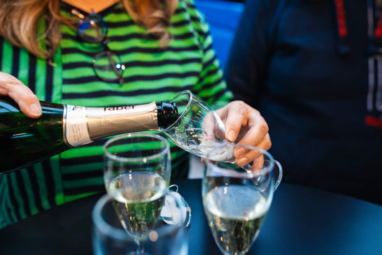 Person in grün-schwarz gestreiftem Pullover gießt Sekt in ein Glas auf einem Tisch mit mehreren gefüllten Gläsern.