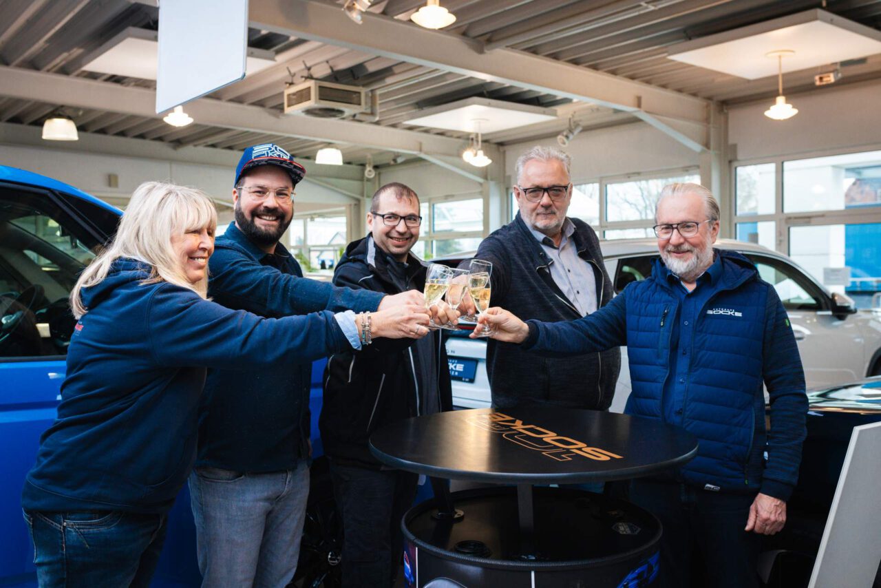Mehrere Personen stoßen mit Gläsern an, stehen um einen runden Stehtisch in einem Autohaus mit Fahrzeugen im Hintergrund.