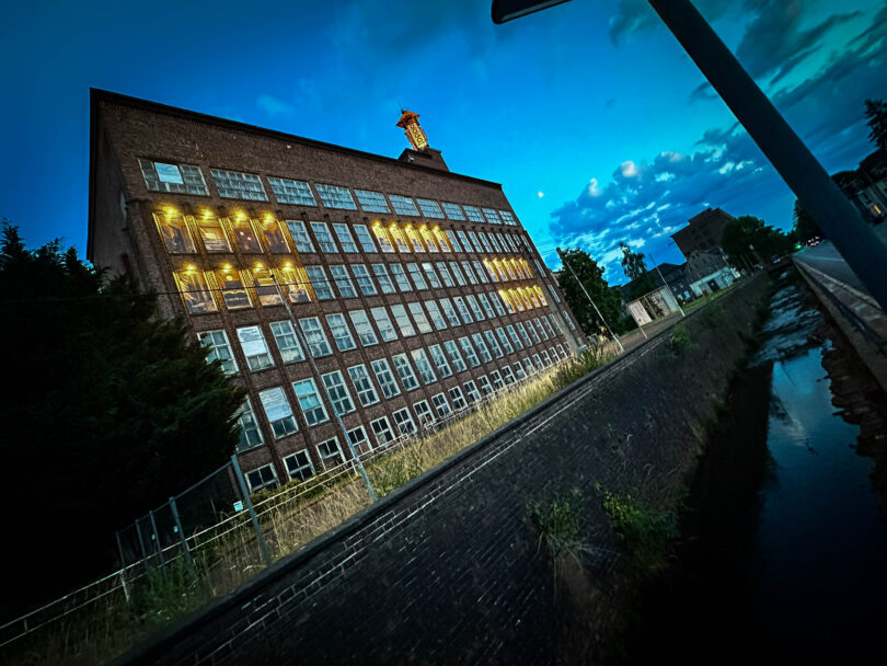 Schräg aufgenommener Backsteinbau mit beleuchteten Fenstern neben einem Wassergraben bei Abenddämmerung