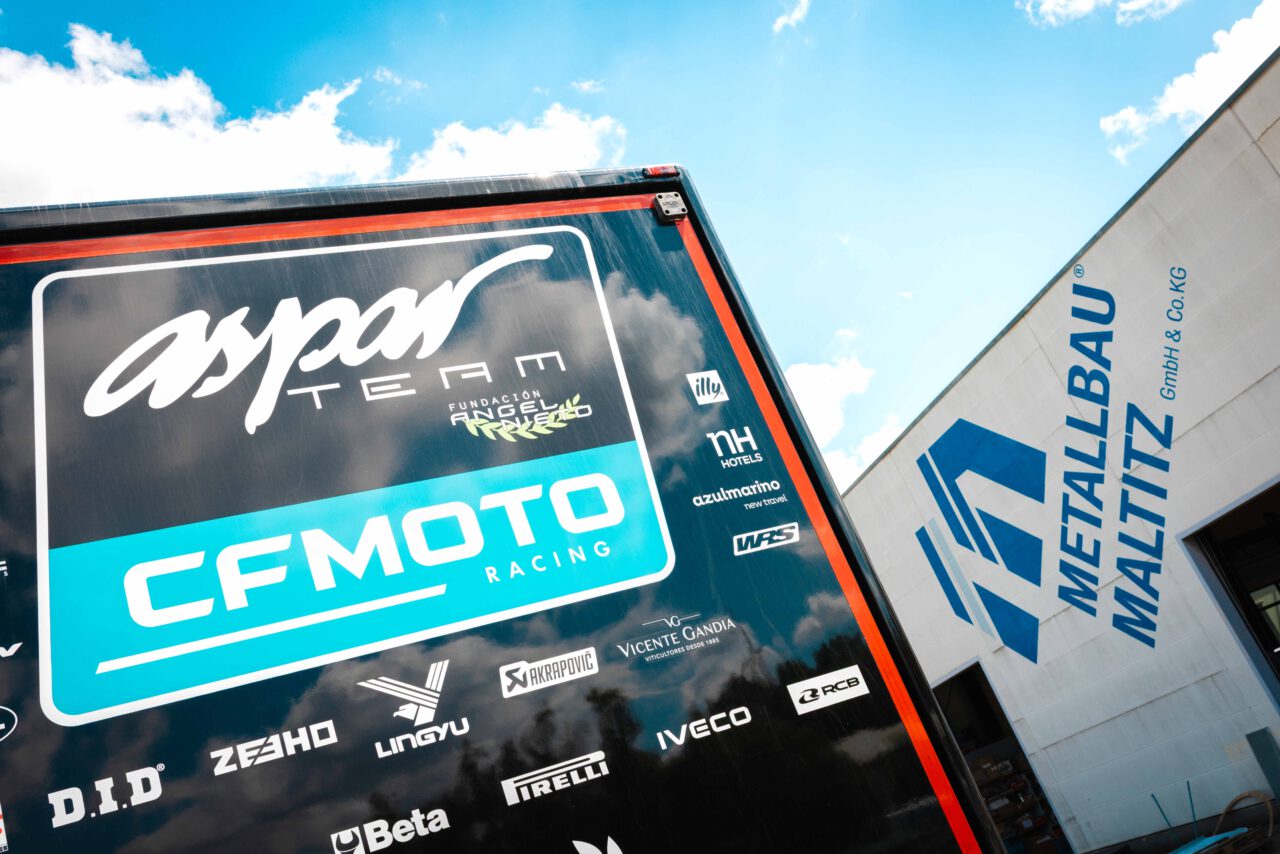 Schwarzer Anhänger mit weißen und blauen Logos von CFMOTO Racing und weiteren Sponsoren, daneben weißes Gebäude mit blauem Schriftzug 'Metallbau Maltitz GmbH & Co. KG'