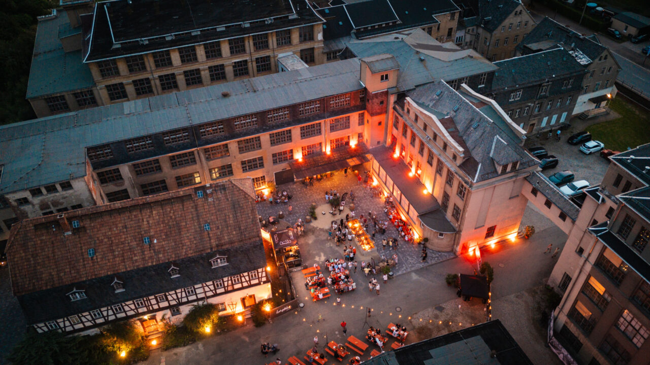 Abendliche Drohnenaufnahme einer Veranstaltung im rustikalen Fabrikhof mit Lichterketten, Fachwerkelementen und stimmungsvoller Beleuchtung.