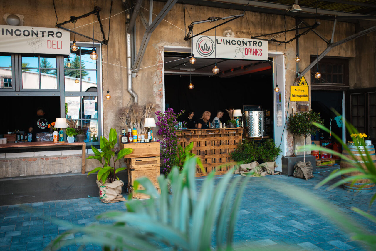 Gastronomie-Außenbereich mit Holz-Theke, Schildern für Drinks und Deli sowie dekorativen Pflanzen vor einer rustikalen Fassade.